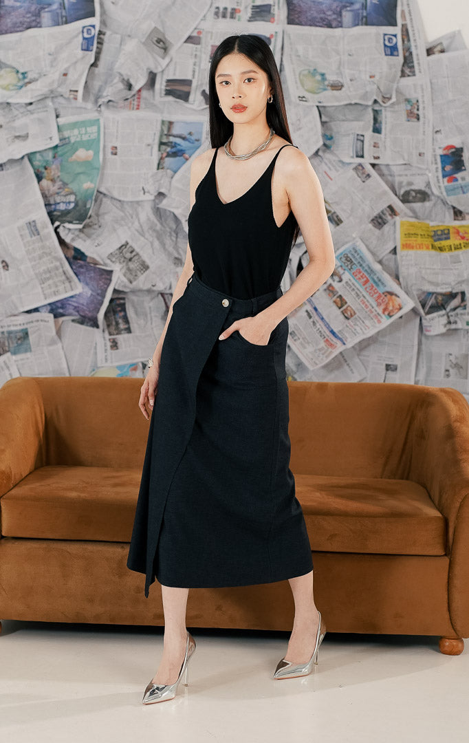 Wanita mengenakan Marjorie Skirt warna navy blue, rok midi panjang dengan model wrap asimetris, detail kancing depan, dan saku samping.
