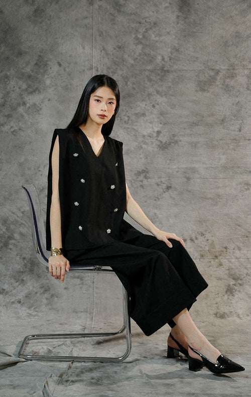 Atasan rompi wanita Marzia warna hitam tanpa lengan dengan potongan leher v-neck, dilengkapi detail aksen payet mewah dan bros bentuk bunga yang manis.