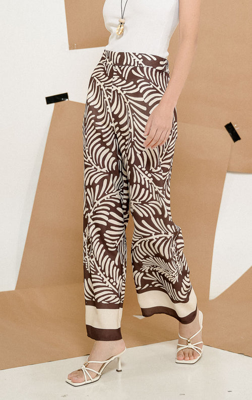 Model mengenakan Mckinney Pants Dark Brown, produk celana panjang kulot wanita potongan longgar warna cokelat tua dengan motif daun botanical tropis.