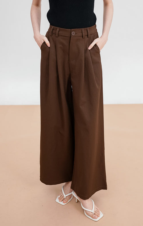 Model mengenakan Micaela Pants warna dark brown, celana kulot wide-leg potongan pinggang tinggi (high-waist) dengan detail lipit depan, kancing senada, dan saku samping.