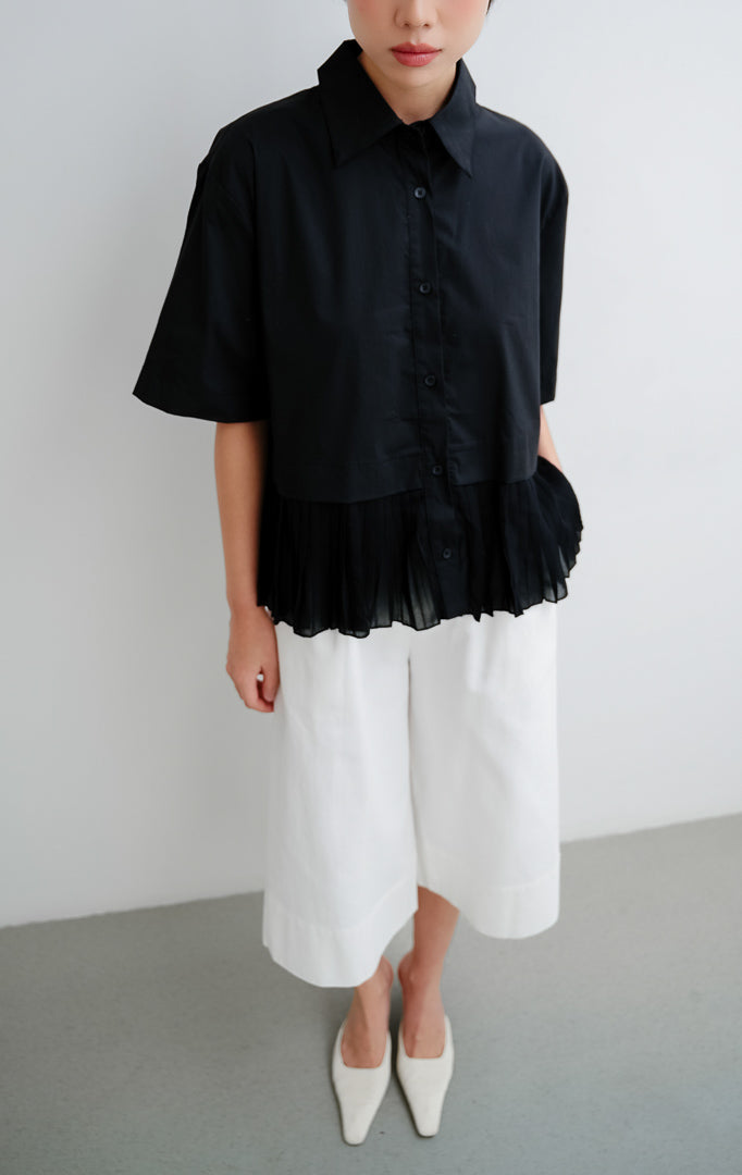 Atasan blouse wanita warna hitam Mildred Top dengan detail lipit (pleats), siluet peplum yang anggun, kancing depan (button-up), dan bahan katun poliester.