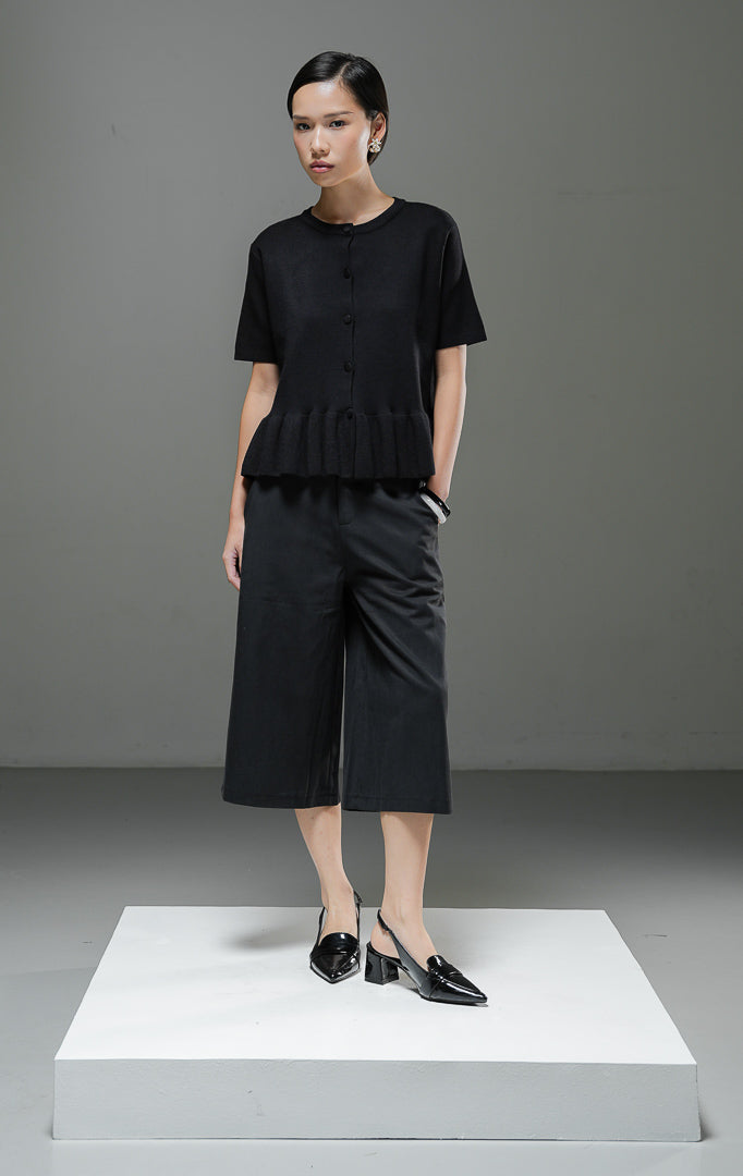 Atasan rajut wanita Naari Top warna hitam lengan pendek dengan kerah bulat, kancing depan bungkus, dan detail rumbai (ruffle hem) rajut di bagian bawah.