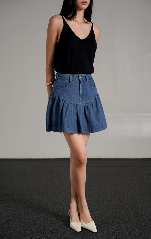 Nathalia Skirt Indigo, rok mini jeans warna biru tua dengan detail lipatan ruffle di bagian bawah, dipadukan dengan tank top hitam.