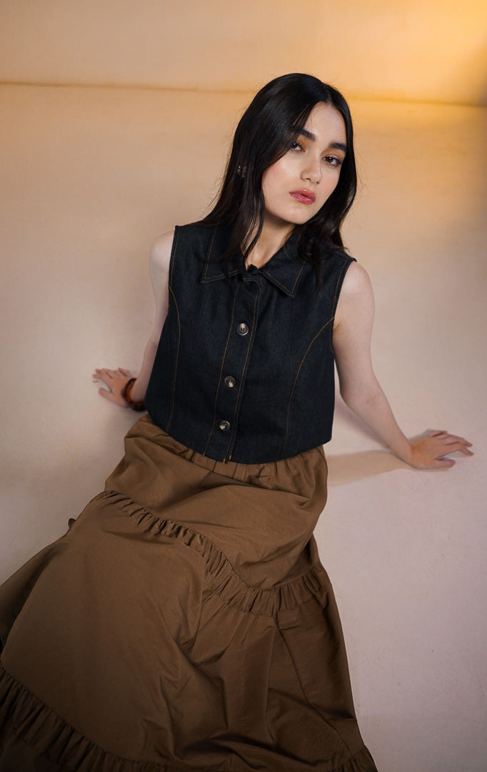 Model wanita mengenakan Norani Sleeveless Crop Denim Top, atasan rompi denim biru tua berkerah dengan kancing depan dan jahitan benang kuning kontras, dipadukan dengan rok rimpel panjang warna cokela