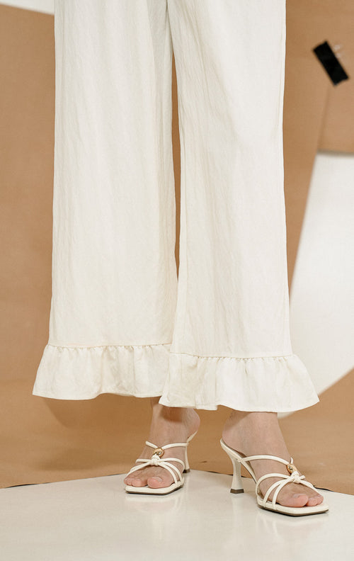 Close-up detail keliman bagian bawah Pericles Pants White, produk celana panjang wanita warna putih dengan aksen ruffle manis di pergelangan kaki.