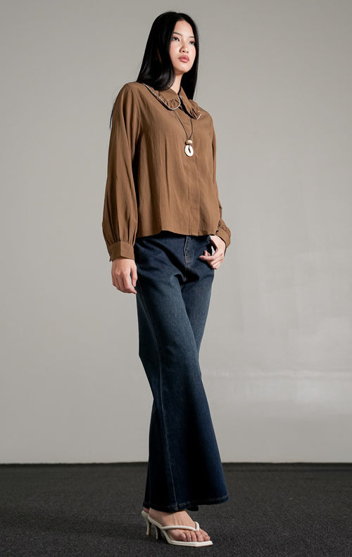 Rebecca Top Brown, kemeja katun lengan panjang warna cokelat dengan detail kerah scallop lebar, dipadukan dengan celana jeans biru tua.