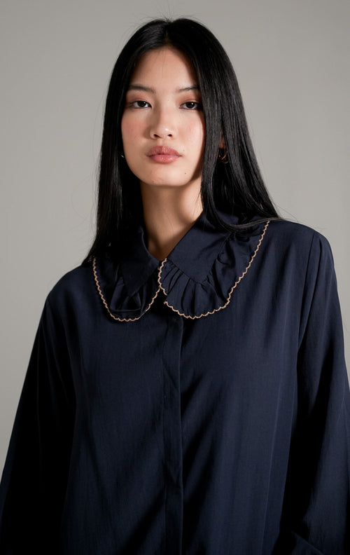Rebecca Top Navy, kemeja katun lengan panjang warna biru tua (navy) dengan kerah scallop beraksen gelombang, dipadukan dengan celana bahan cokelat.