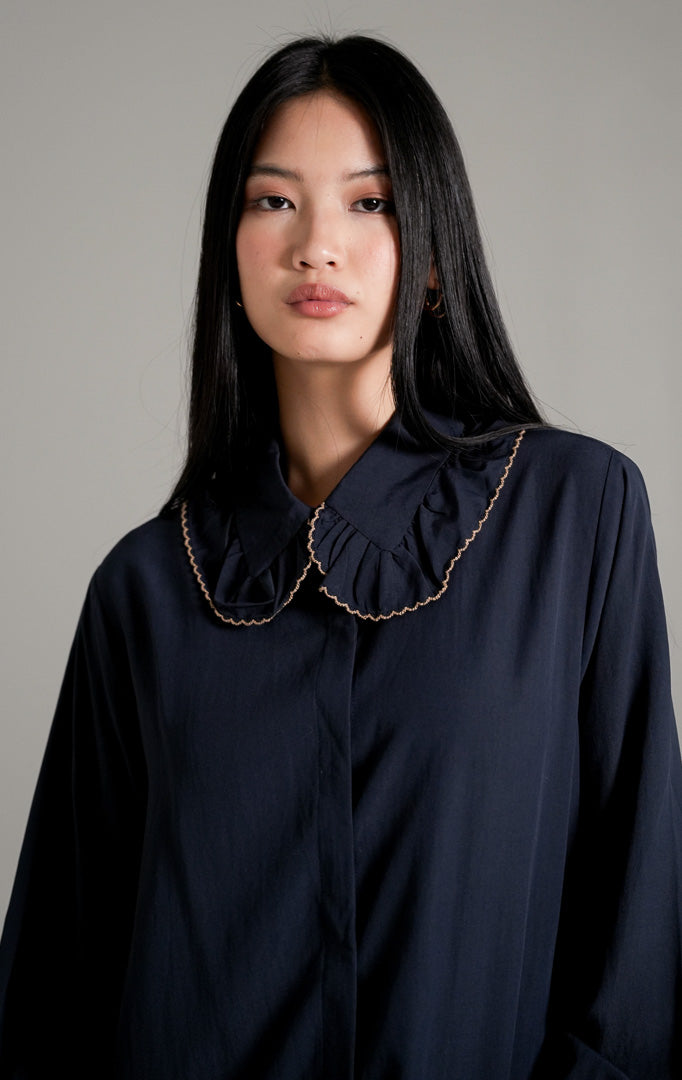 Rebecca Top Navy, kemeja katun lengan panjang warna biru tua (navy) dengan kerah scallop beraksen gelombang, dipadukan dengan celana bahan cokelat.