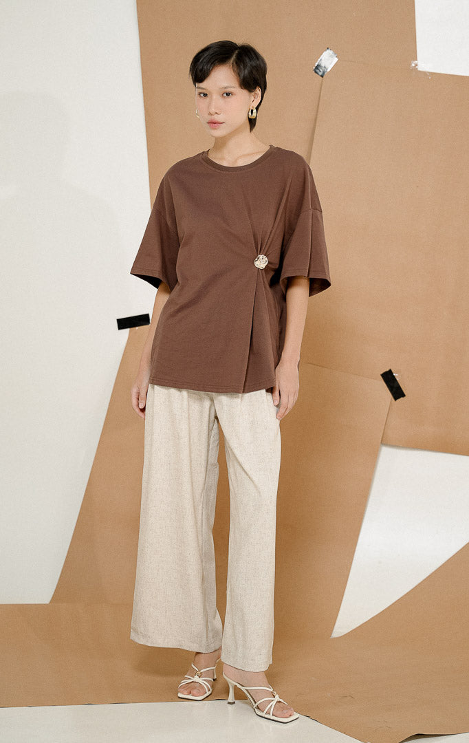 Close up model mengenakan Greta Short Sleeved Top Brown, produk atasan kaos wanita lengan pendek warna cokelat dengan aksen bros emas lepasan di samping.