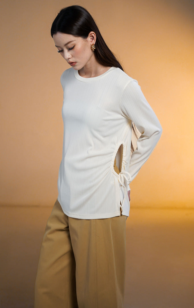 Model wanita mengenakan Rinata Long Sleeve Top warna putih gading (ivory), atasan lengan panjang bertekstur rib halus dengan detail tali serut (drawstring) dan lubang di sisi pinggang kiri, dipadukan dengan celana panjang cokelat mustard