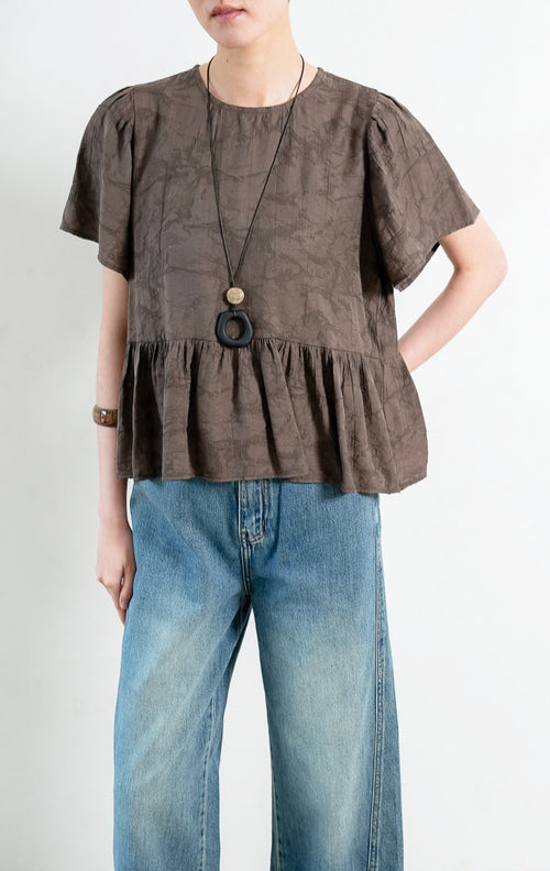 Atasan wanita lengan pendek warna cokelat tua (dark brown) Rivena Top berbahan tekstur crinkle ringan (lightweight crinkle texture) dengan kerah bulat (round neckline) dan detail bagian bawah bertingkat kerut (softly gathered tiered hem).