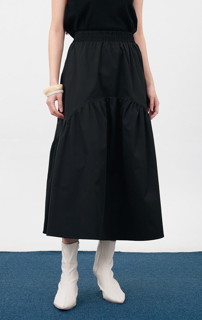 Model mengenakan Rosalia Skirt warna black, rok panjang dengan detail pinggang elastis dan potongan bertingkat (tiered) yang melebar di bagian bawah.