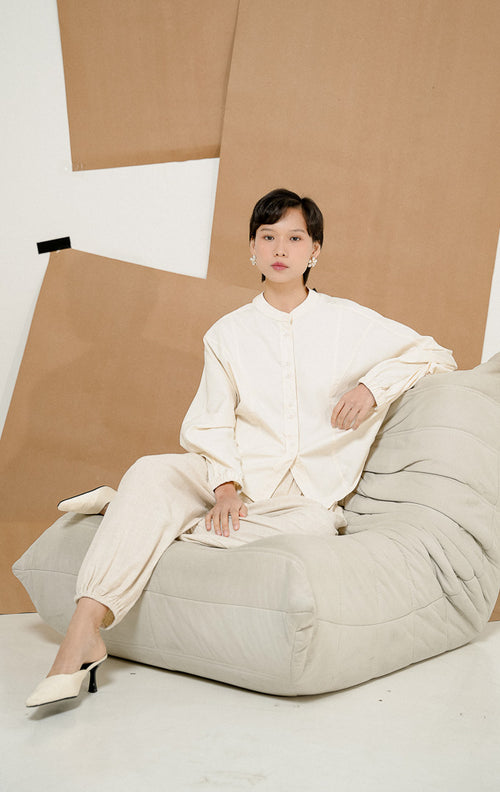 Model duduk di sofa beanbag mengenakan Rosemarie Top Ivory, produk atasan blouse kemeja wanita lengan panjang warna putih gading dengan detail kancing depan.