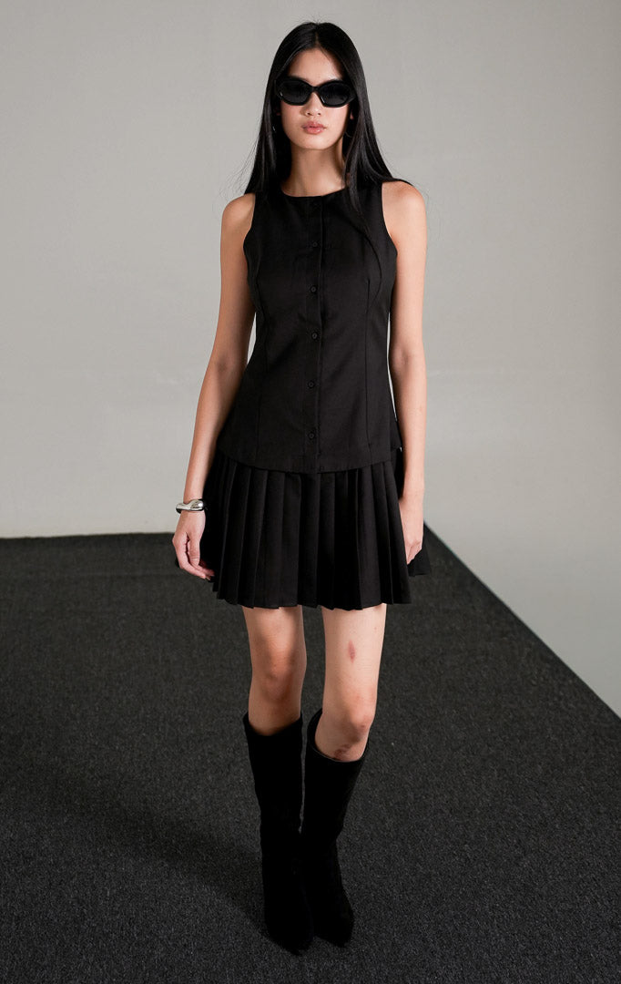 Rosemary Dress Black, gaun mini tanpa lengan warna hitam dengan kancing depan dan rok lipit pendek, dipadukan dengan kacamata hitam dan sepatu boots tinggi.
