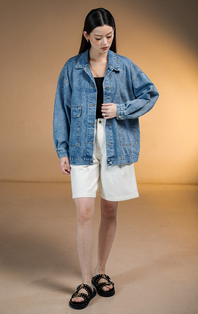 Model wanita memakai Rowey Jacket Denim, jaket jeans oversized warna biru muda dengan saku depan besar dan kancing perak, dipadukan dengan celana pendek putih