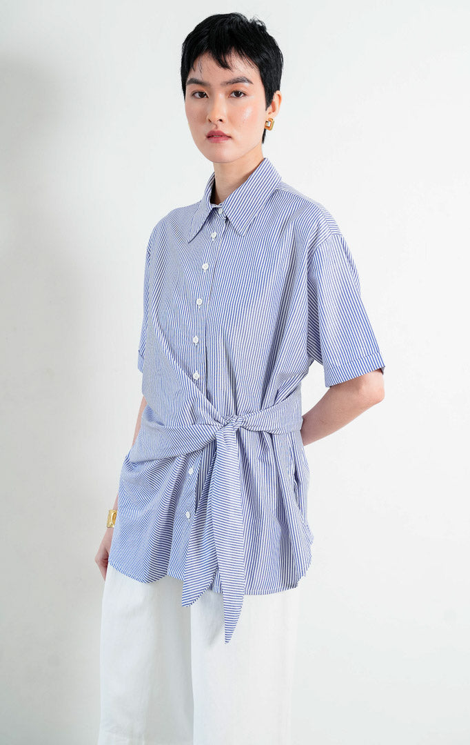 Kemeja wanita lengan pendek motif salur biru putih (blue and white pinstripes) Sacha Top dengan kerah kemeja (pointed collar), bukaan kancing depan, detail tali ikat di samping (side tie details at hem), dan kelim melengkung (curved hem).