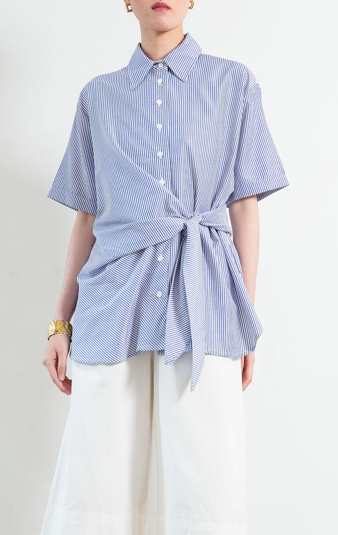 Kemeja wanita lengan pendek motif salur biru putih (blue and white pinstripes) Sacha Top dengan kerah kemeja (pointed collar), bukaan kancing depan, detail tali ikat di samping (side tie details at hem), dan kelim melengkung (curved hem).