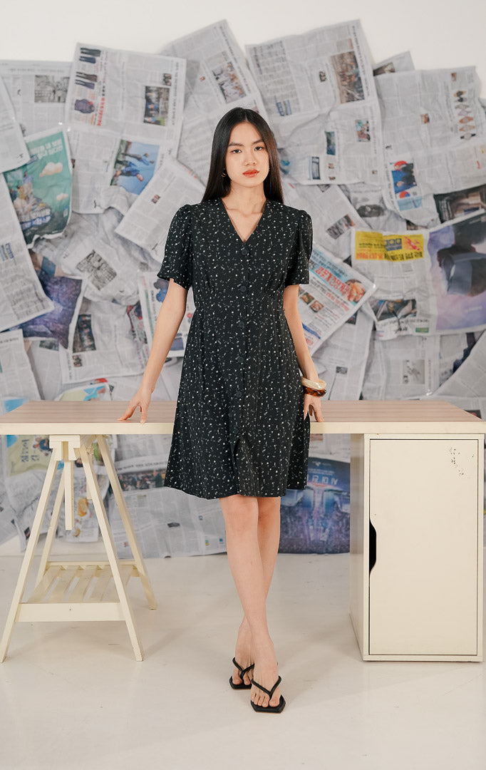Model mengenakan Seraphina Dress Black, dress wanita warna hitam dengan motif floral kecil yang anggun, lengan pendek, dan potongan feminin yang nyaman.
