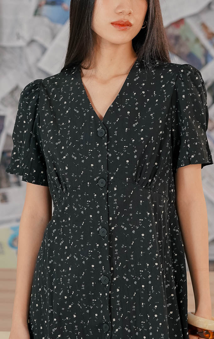 Model mengenakan Seraphina Dress Black, dress wanita warna hitam dengan motif floral kecil yang anggun, lengan pendek, dan potongan feminin yang nyaman.