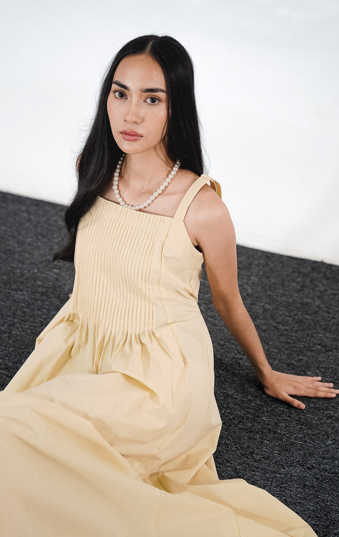 Skylar Dress warna kuning, gaun A-line tanpa lengan dengan detail lipatan pintuck di bagian dada dan saku samping, dipadukan dengan kalung mutiara