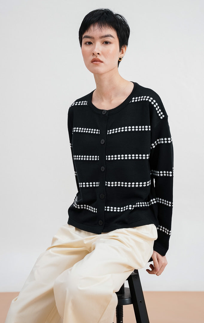 Model mengenakan Soleil Cardigan warna black, kardigan rajut lengan panjang dengan motif garis horizontal titik-titik putih dan kancing bulat hitam di bagian depan.