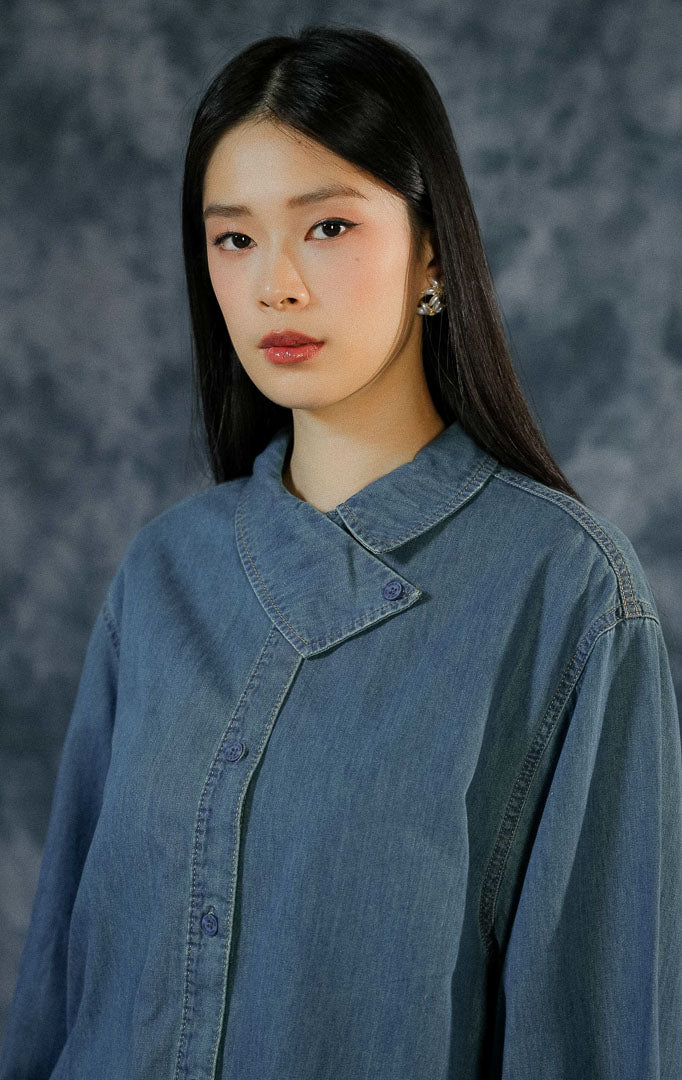 Kemeja lengan panjang wanita Steya berbahan denim dengan detail kerah asimetris yang unik (statement collar), kancing depan aktif, dan lengan kancing.