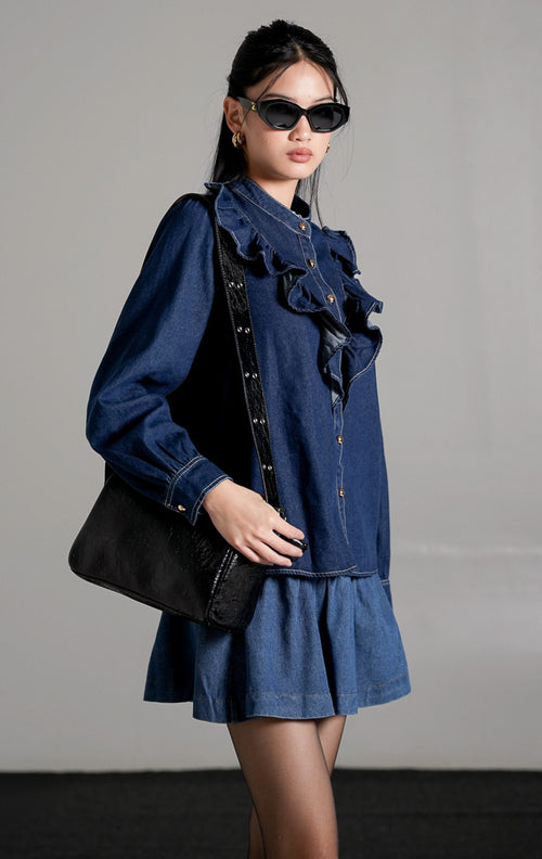 Sylvain Top Dark Denim, kemeja denim biru tua dengan detail ruffle asimetris di bagian dada dan kancing emas, dipadukan dengan rok mini denim senada