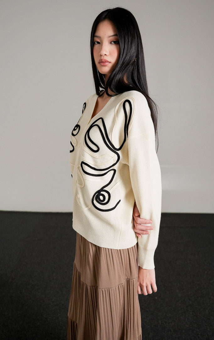 Thea Patterned Knit Cardigan, kardigan rajut warna broken white dengan motif garis melengkung hitam, dipadukan dengan rok panjang cokelat.