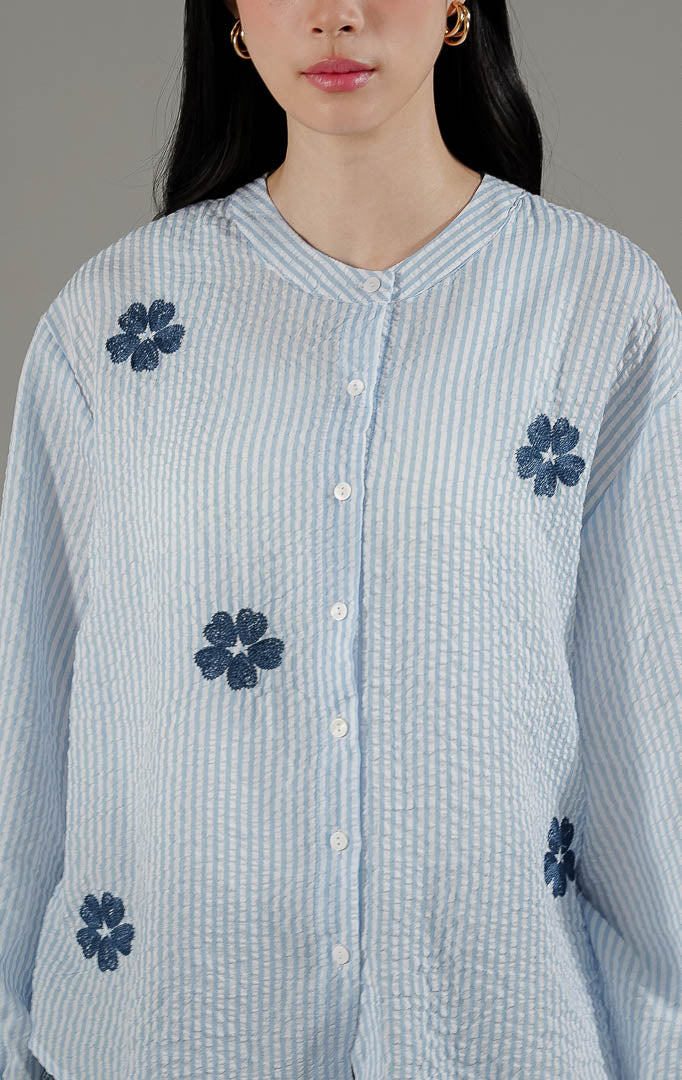 Azriel Top Blue dari This Is April, kemeja stripe biru putih lengan panjang dengan detail bordir bunga hitam dan pergelangan tangan karet.