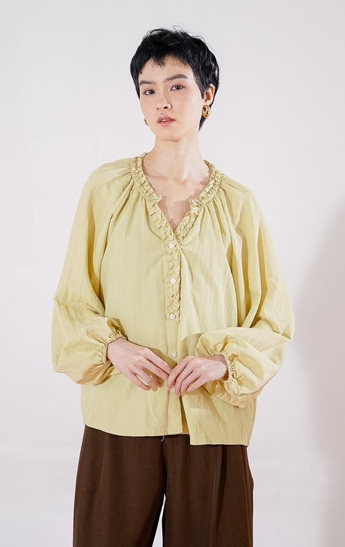 Blouse lengan panjang wanita This Is April Eloise Top warna khaki berbahan katun, dengan kerah V-neck beraksen ruffle, kancing depan, dan ujung lengan karet.