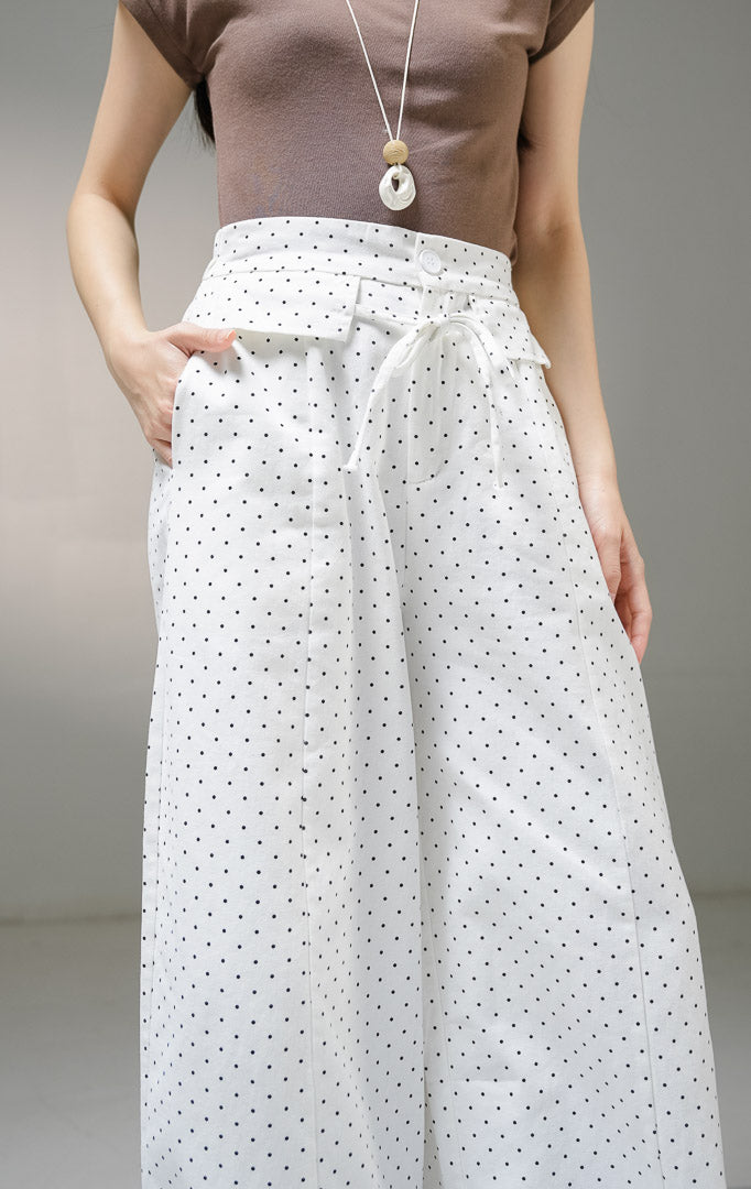 Celana panjang kulot wanita This Is April Evelyna Pants warna putih motif polkadot hitam kecil, dilengkapi kancing depan, saku penutup dekoratif, dan tali ikat pinggang.