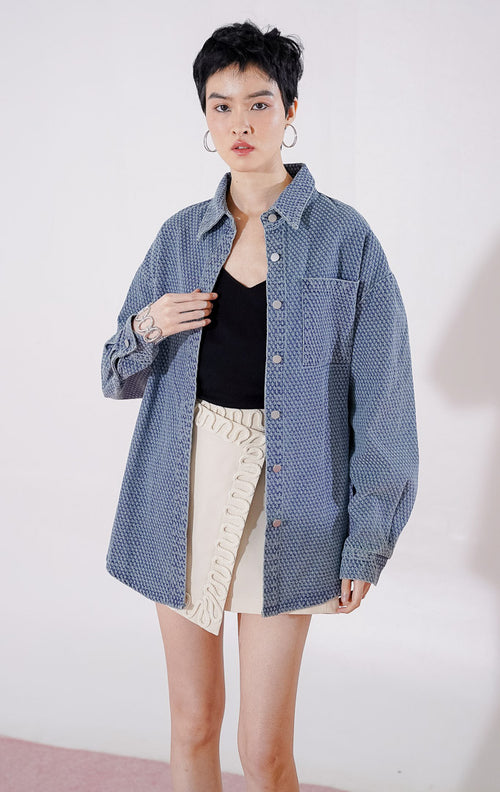 Kemeja wanita lengan panjang Knox Top Denim dari This Is April berwarna biru dengan detail tekstur topstitching di seluruh bagian, dilengkapi kantong dada dan kancing depan, digantung di rak pakaian.