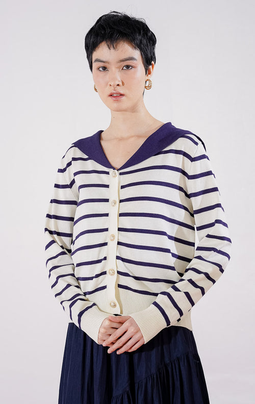 Cardigan rajut wanita lengan panjang Leone Cardigan dari This Is April berwarna ivory dengan motif garis horizontal biru navy, dilengkapi kerah lebar berwarna solid navy dan kancing emas di bagian depan, digantung di rak pakaian.