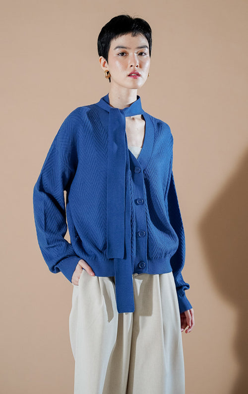 Marianne Cardigan Blue dari This Is April, cardigan rajut V-neck warna biru dengan kancing depan dan detail syal kain yang menyatu di bagian leher.