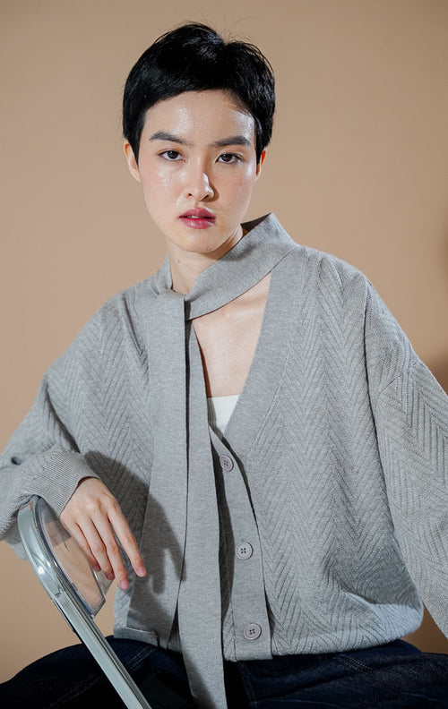 Model wanita mengenakan This Is April Marianne Cardigan Grey, kardigan rajut V-neck abu-abu dengan kancing depan, detail syal leher menyatu, dan motif rajutan chevron.