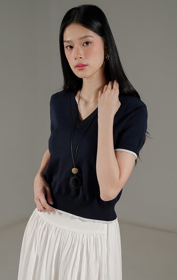 Morgane Top Navy dari This Is April, atasan rajut V-neck lengan pendek warna biru navy dengan detail garis putih kontras pada ujung lengan dan pinggang.