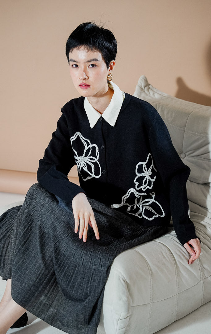 Peppie Cardigan Black dari This Is April, cardigan rajut warna hitam dengan detail bordir bunga putih di bagian depan dan kancing aktif.