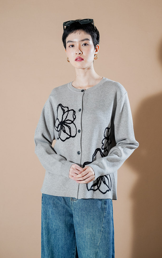 Peppie Cardigan Grey dari This Is April, cardigan rajut warna abu-abu dengan detail bordir bunga hitam di sisi depan dan kancing aktif.