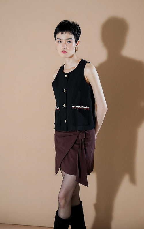 Model wanita mengenakan This Is April Shailene Vest Black, rompi hitam tanpa lengan dengan kancing depan, dipadukan dengan rok celana cokelat.