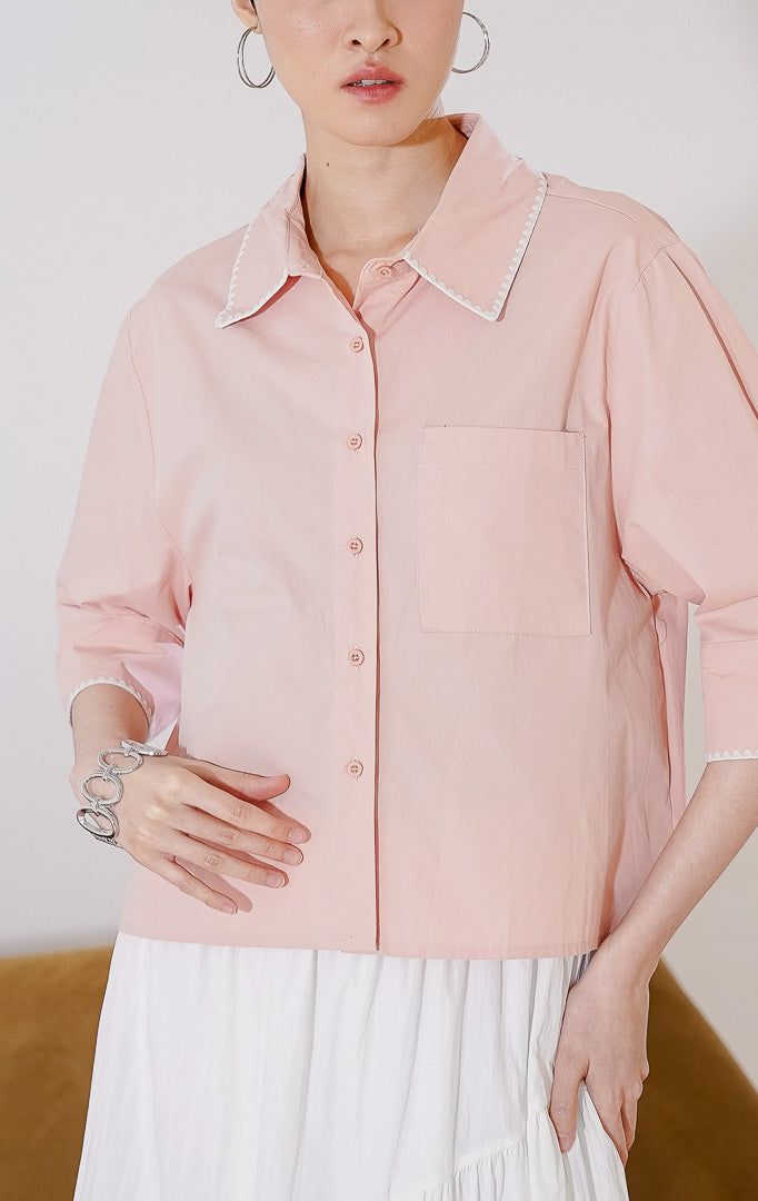 Kemeja wanita lengan 3/4 Venus Top dari This Is April berwarna dusty pink. Dilengkapi detail jahitan zigzag putih di tepi kerah dan ujung lengan, saku dada kiri fungsional, dan kancing depan, digantung di rak pakaian.