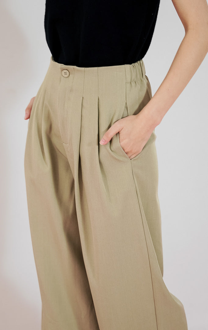 Model wanita mengenakan celana panjang This Is April Aeron Pants warna sage green berpotongan straight-leg dengan detail pleat depan, dipadukan dengan tanktop hitam.