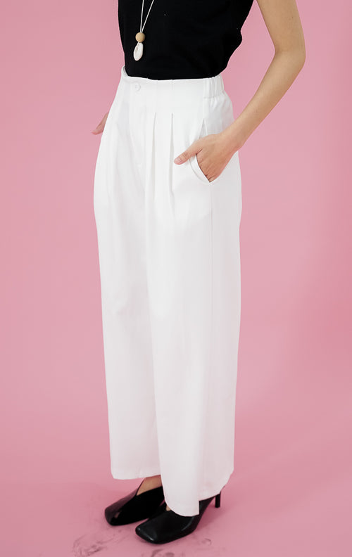 Model wanita mengenakan celana panjang This Is April Aeron Pants warna putih berpotongan straight-leg dengan detail pleat depan, dipadukan dengan tanktop hitam dan kalung panjang.