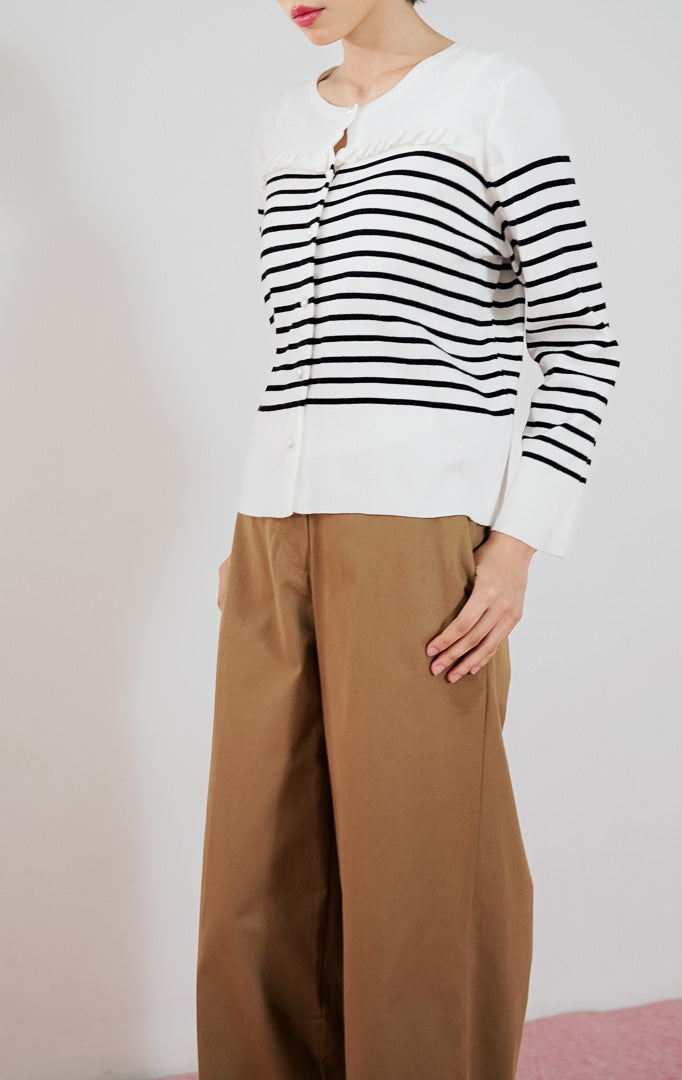 Model wanita mengenakan cardigan rajut lengan panjang This Is April Alexiz Cardigan warna putih motif garis hitam dengan kancing depan dan detail frill, dipadukan dengan celana panjang cokelat dan sepatu boots putih.