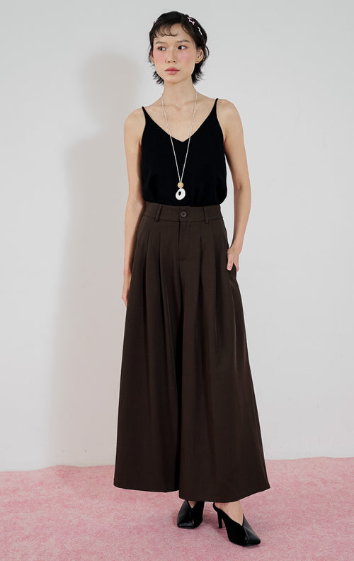 Model wanita mengenakan celana kulot berpotongan lebar This Is April Ally Pants warna dark brown dengan detail pleat depan, dipadukan dengan tanktop v-neck hitam, kalung panjang, dan sepatu mules hitam.