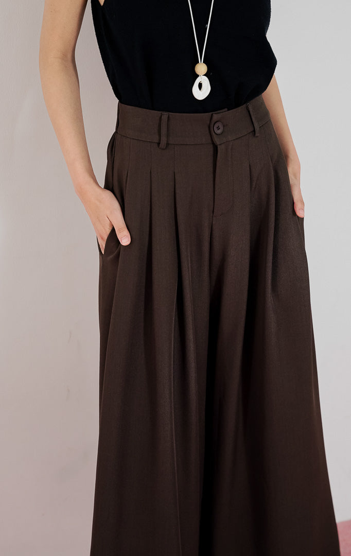 Model wanita mengenakan celana kulot berpotongan lebar This Is April Ally Pants warna dark brown dengan detail pleat depan, dipadukan dengan tanktop v-neck hitam, kalung panjang, dan sepatu mules hitam.