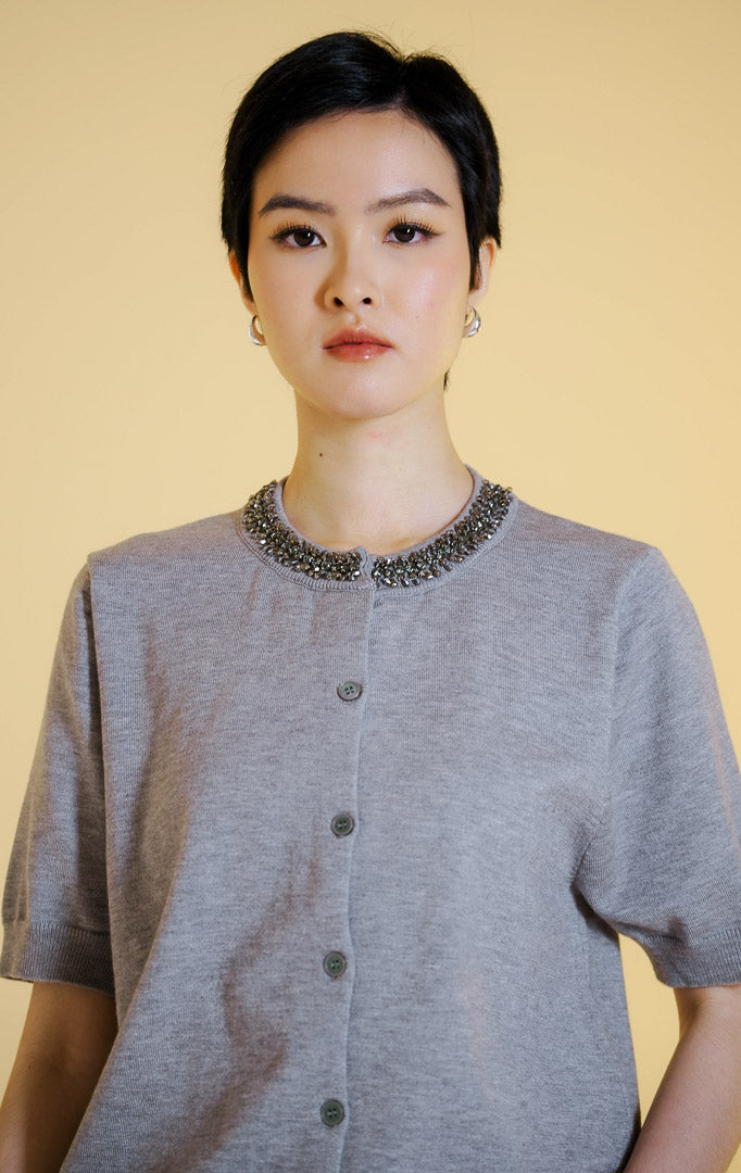 Pakaian wanita model atasan rajut cardigan lengan pendek dengan kancing depan dan hiasan permata di bagian kerah bulat (gemstone neckline short sleeve knit top) warna abu-abu muda Alveria Top untuk gaya kantor dan kasual anggun.