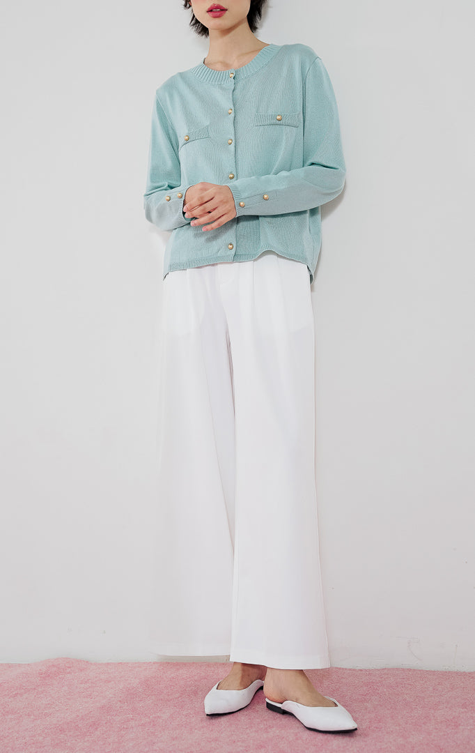 Model wanita mengenakan cardigan rajut klasik lengan panjang This Is April Arnon Cardigan warna tosca dengan kancing emas dan aksen saku, dipadukan dengan celana panjang kulot putih dan sepatu flat putih.