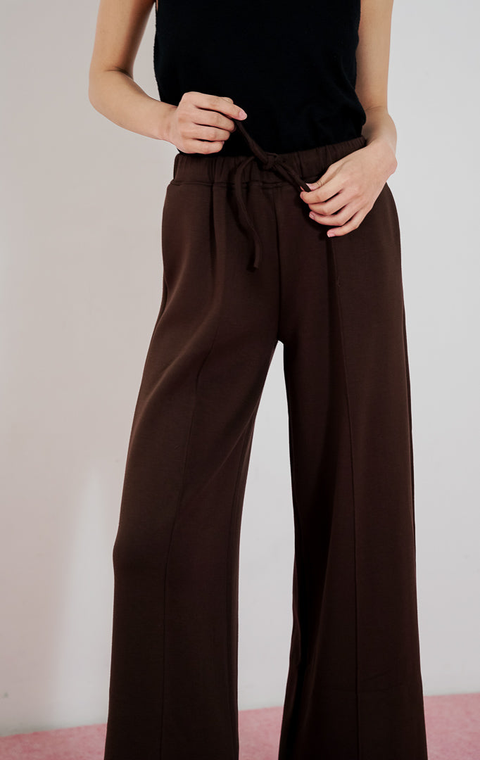 Model wanita mengenakan celana panjang This Is April Cora Pants warna dark brown berpotongan straight-leg dengan detail tali serut di pinggang, dipadukan dengan tanktop hitam.
