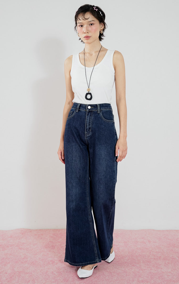 Model mengenakan celana jeans wanita This Is April Dolores warna dark denim berpotongan wide-leg, dipadukan dengan tanktop putih dan kalung aksesoris.