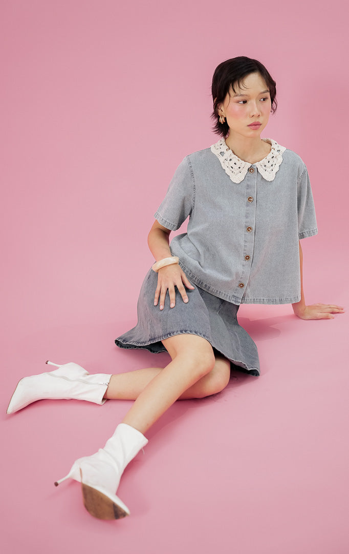 Model mengenakan This Is April Elliana Top, kemeja denim lengan pendek warna light denim dengan detail kerah renda crochet putih, dipadukan skort denim senada.